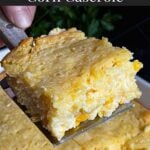 A serving of creamy corn casserole lifted over the baking pan on a dark surface.