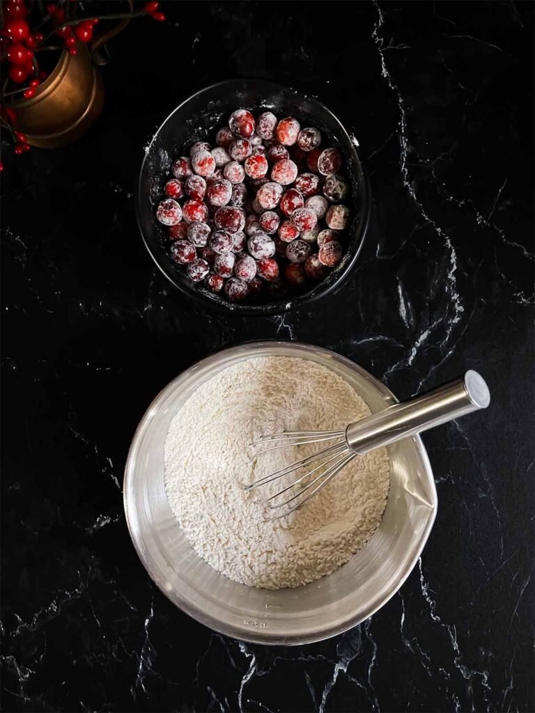 A bowl of sugar dusted cranberries and a bowl of a flour mixture on a dark surface.