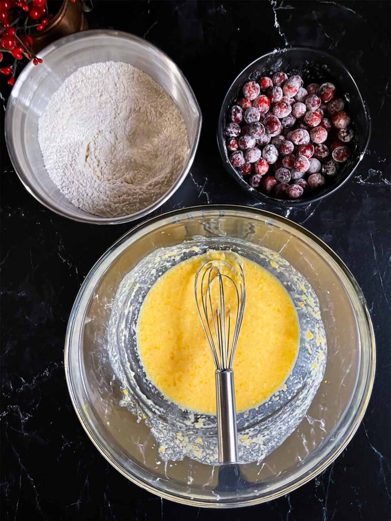 Wet and dry ingredients for Cranberry Orange bread in various bowls on a dark surface.