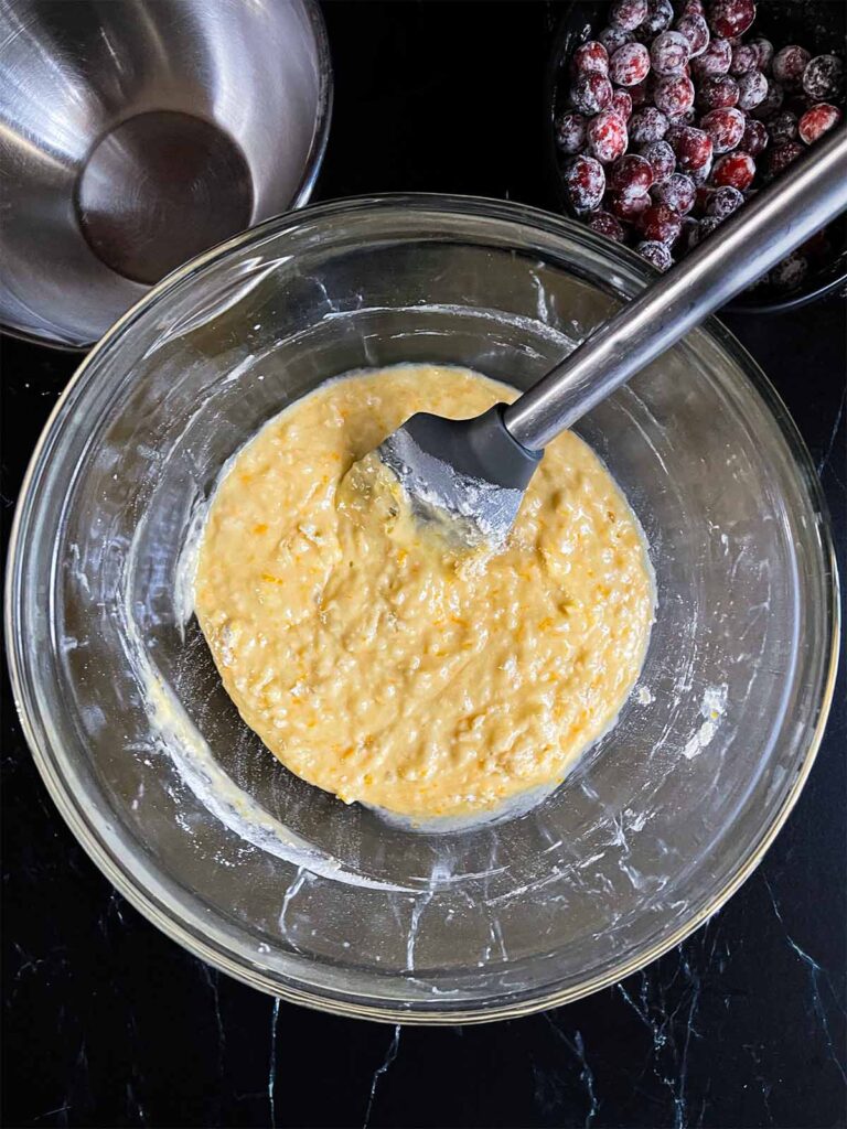 Wet and dry ingredients combined in a glass mixing bowl, with a bowl of cranberries in the background.