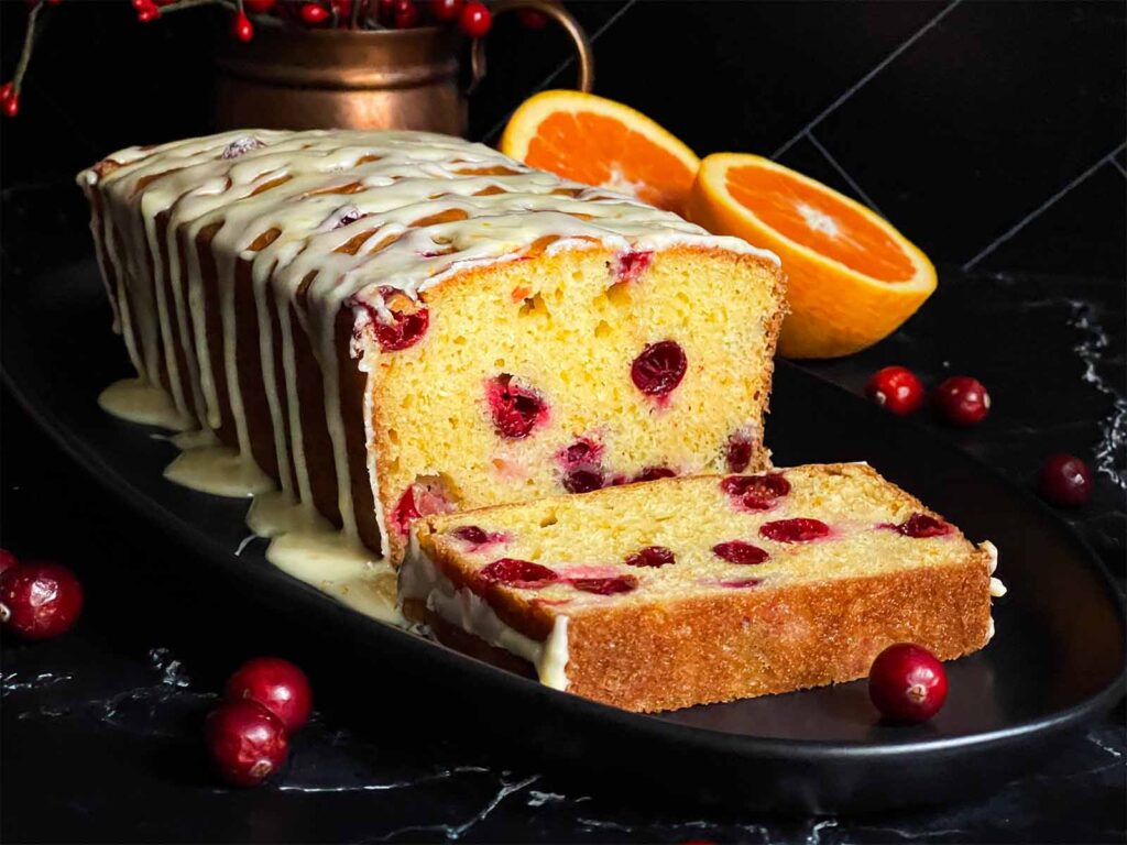 Cranberry Orange bread with an orange glaze on a dark surface with one slice laying on its side showing the interior.