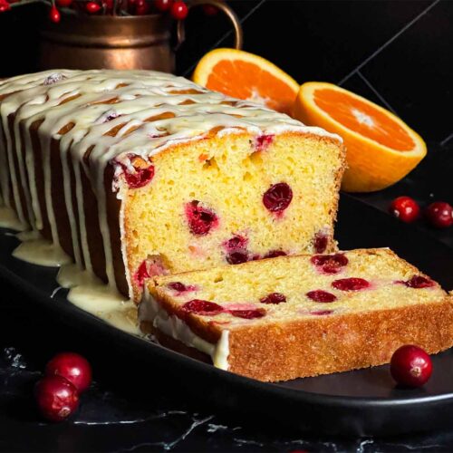 Cranberry Orange bread with an orange glaze on a dark surface with one slice laying on its side showing the interior.