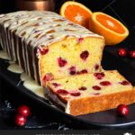 Cranberry Orange bread with an orange glaze on a dark surface with one slice laying on its side showing the interior.