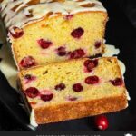 Cranberry Orange bread with an orange glaze on a dark surface with one slice laying on its side showing the interior.