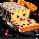 Cranberry Orange bread with an orange glaze on a dark surface with one slice laying on its side showing the interior.