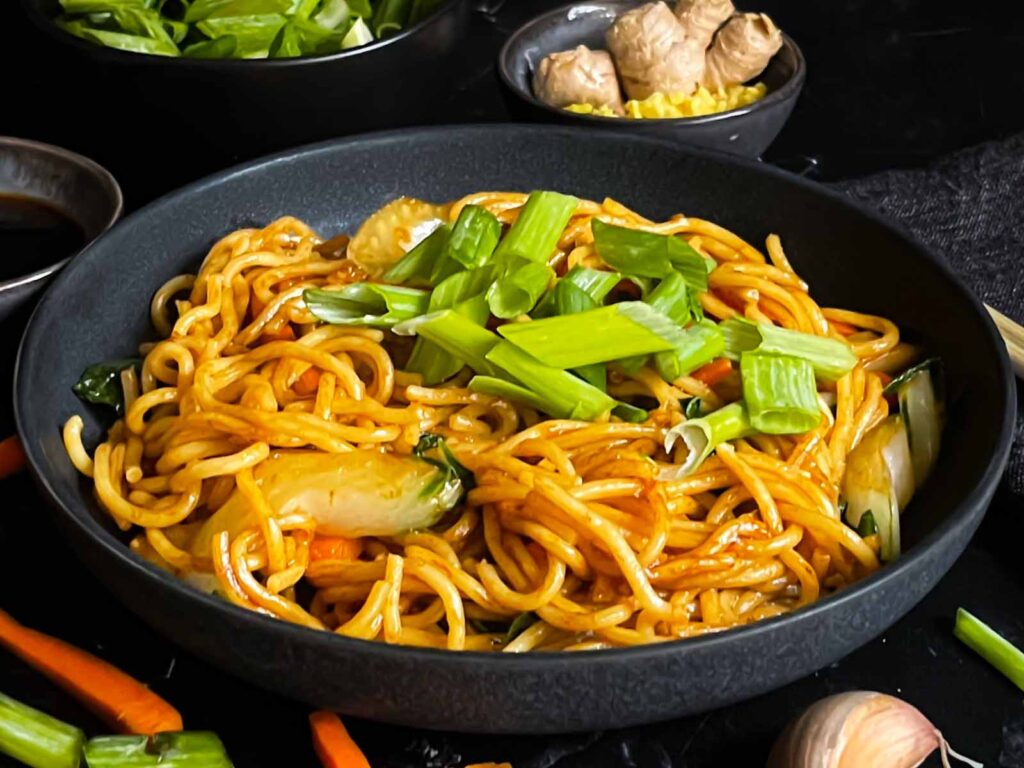 Vegetable lo mein garnished with green onion in a dark bowl.