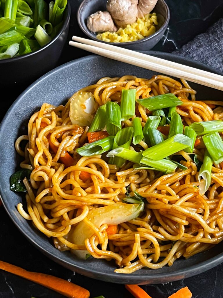 Vegetable lo mein garnished with green onion in a dark bowl.