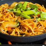 Vegetable lo mein garnished with green onion in a dark bowl.