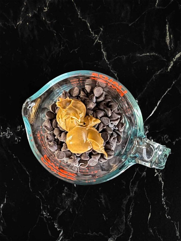 Peanut butter and chocolate chips in a small glass measuring cup on a dark surface.