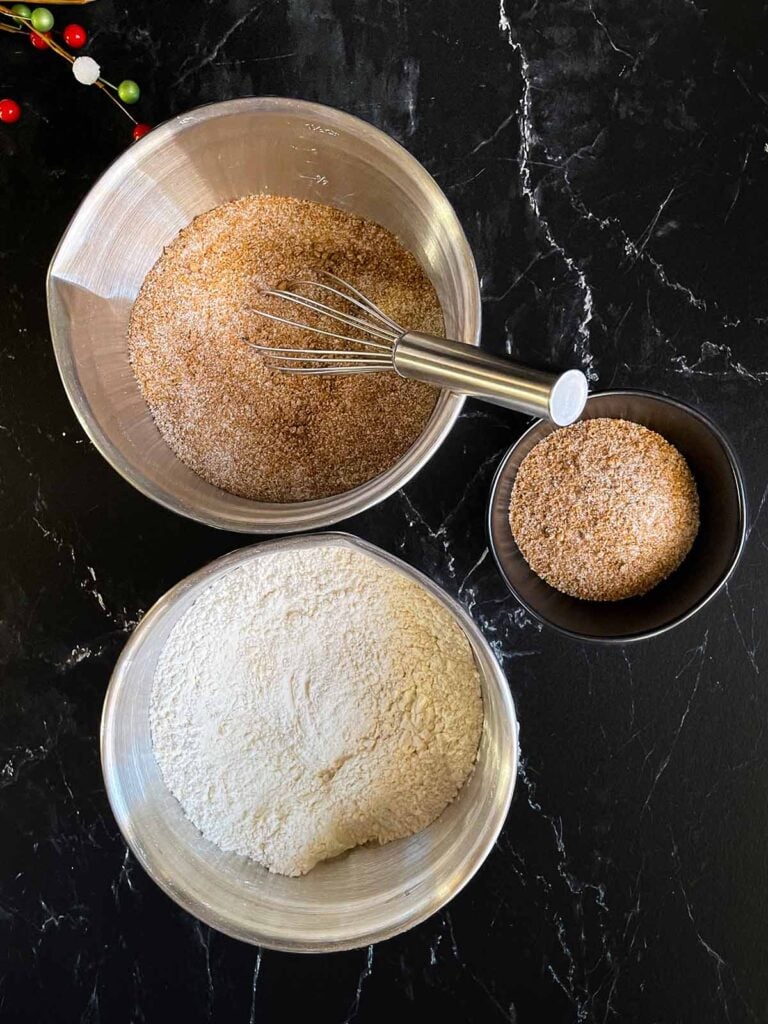Chai sugar mixture in one metal bowl and dry ingredients in another metal bowl on a dark surface.