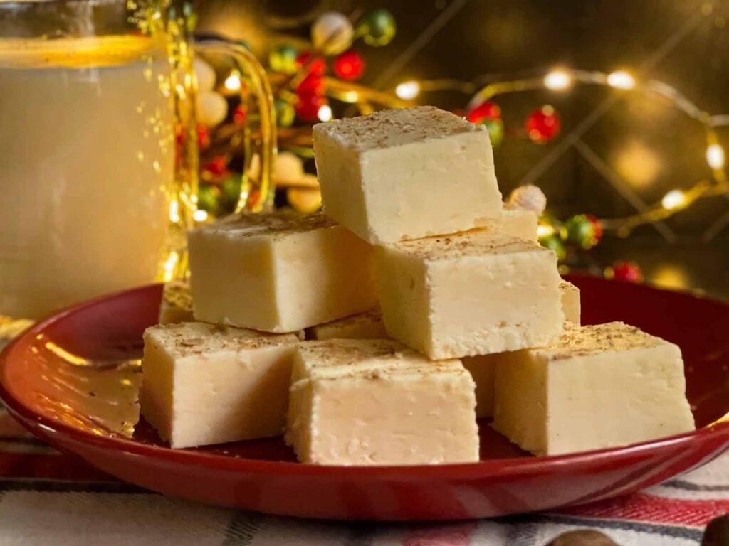 Eggnog fudge stacked on a dark plate with a glass of eggnog.