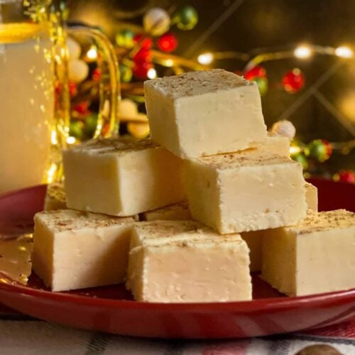 Eggnog fudge stacked on a dark plate with a glass of eggnog.