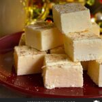 Eggnog fudge stacked on a dark plate with a glass of eggnog.