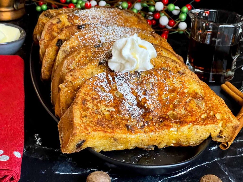 Slices of panettone French toast garnished with powdered sugar, whipped cream, and maple syrup on a dark plate.