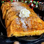 Slices of panettone French toast garnished with powdered sugar, whipped cream, and maple syrup on a dark plate.