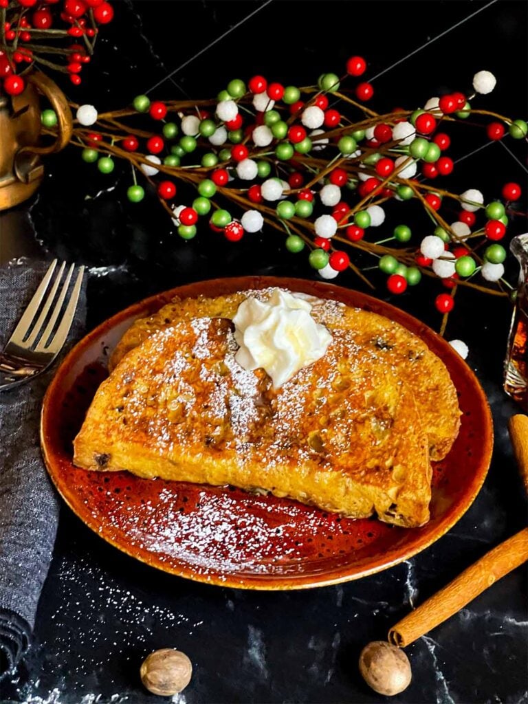 Slices of panettone French toast garnished with powdered sugar, whipped cream, and maple syrup on a dark plate.