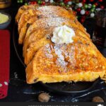 Slices of panettone French toast garnished with powdered sugar, whipped cream, and maple syrup on a dark plate.