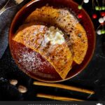 Slices of panettone French toast garnished with powdered sugar, whipped cream, and maple syrup on a red plate.