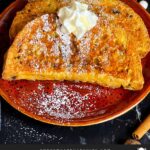 Slices of panettone French toast garnished with powdered sugar, whipped cream, and maple syrup on a red plate.