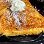Slices of panettone French toast garnished with powdered sugar, whipped cream, and maple syrup on a dark plate.