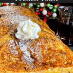 Slices of panettone French toast garnished with powdered sugar, whipped cream, and maple syrup on a dark plate.