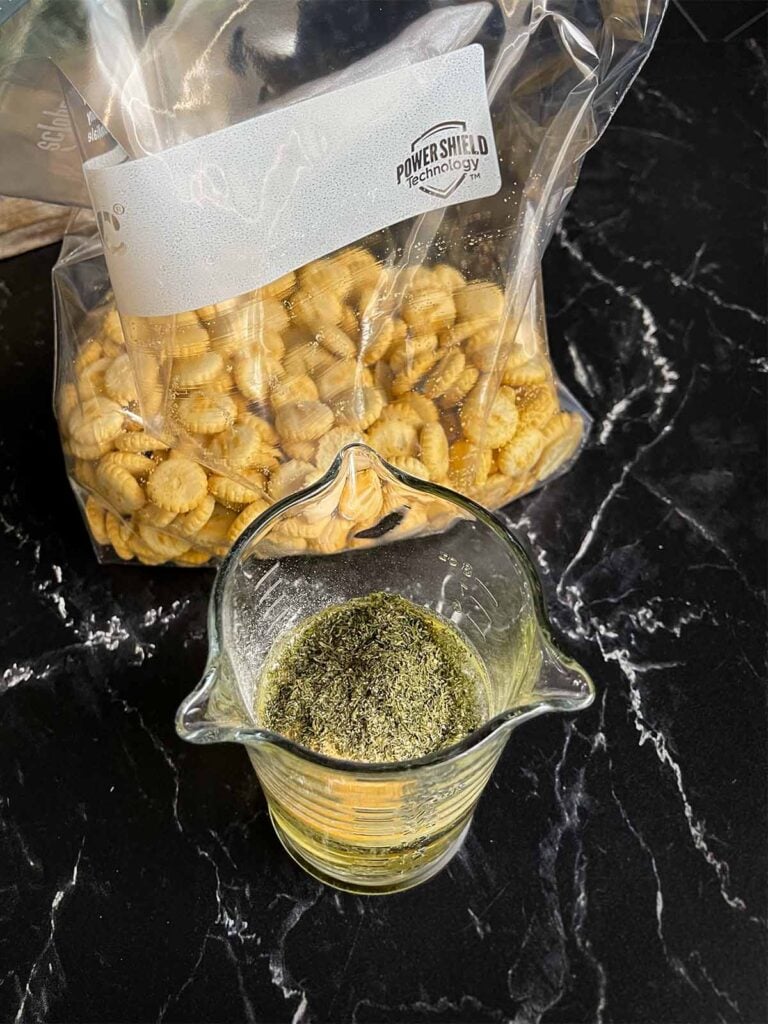 Seasonings for dill oyster crackers in a glass measuring cup on a dark surface.