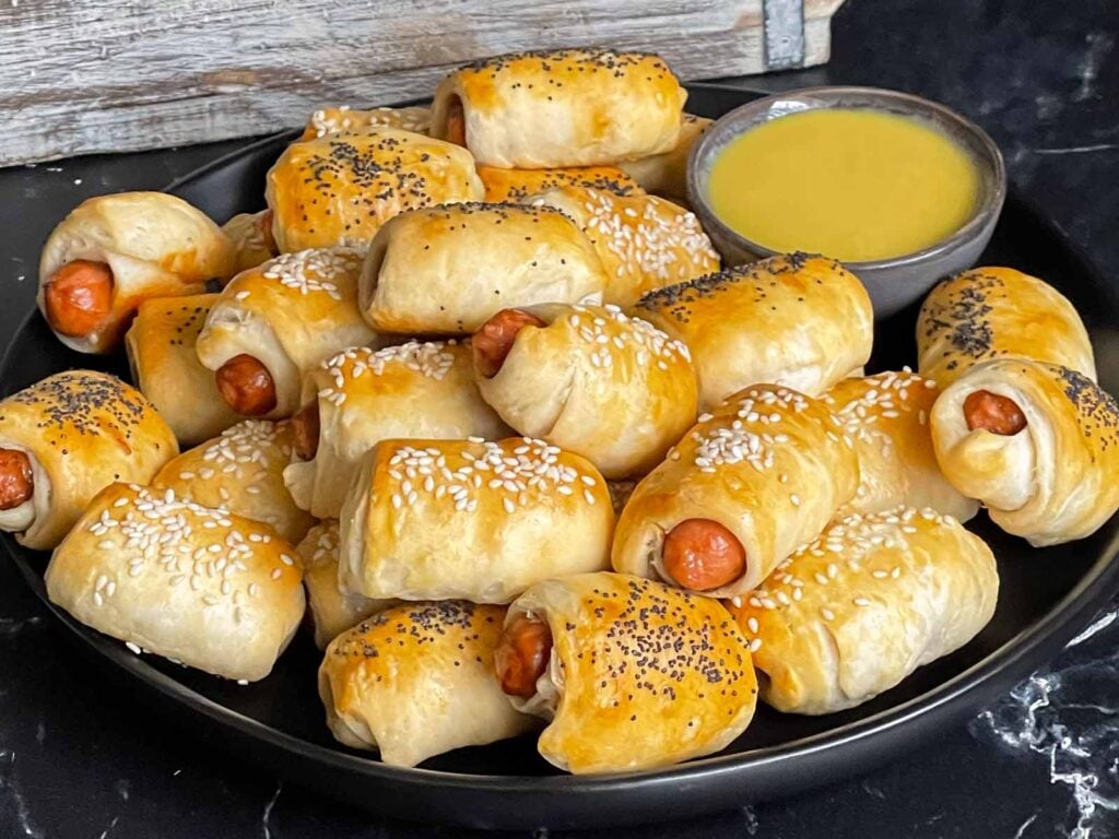 Pigs in a blanket stacked on a dark plate with a ramekin of honey mustard sauce.