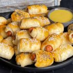 Pigs in a blanket stacked on a dark plate with a ramekin of honey mustard sauce.