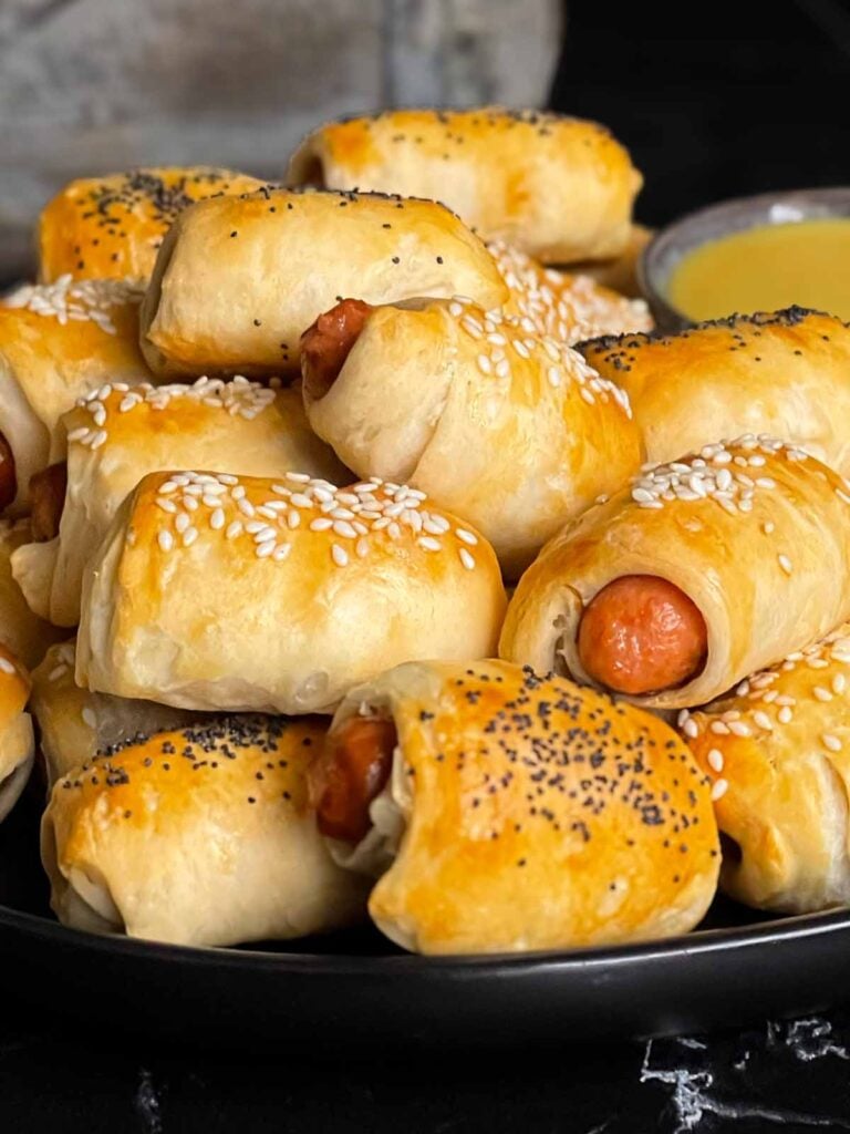 Pigs in a blanket stacked on a dark plate with a ramekin of honey mustard sauce.