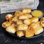 Pigs in a blanket stacked on a dark plate with a ramekin of honey mustard sauce.