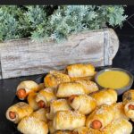 Pigs in a blanket stacked on a dark plate with a ramekin of honey mustard sauce.