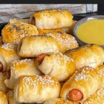 Pigs in a blanket stacked on a dark plate with a ramekin of honey mustard sauce.