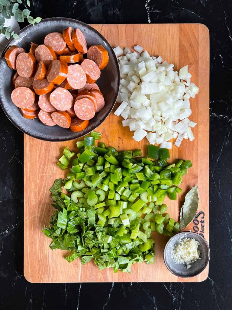 Sliced andouille, onions, green bell pepper, celery, minced garlic and bay leave on a wooden cutting board.
