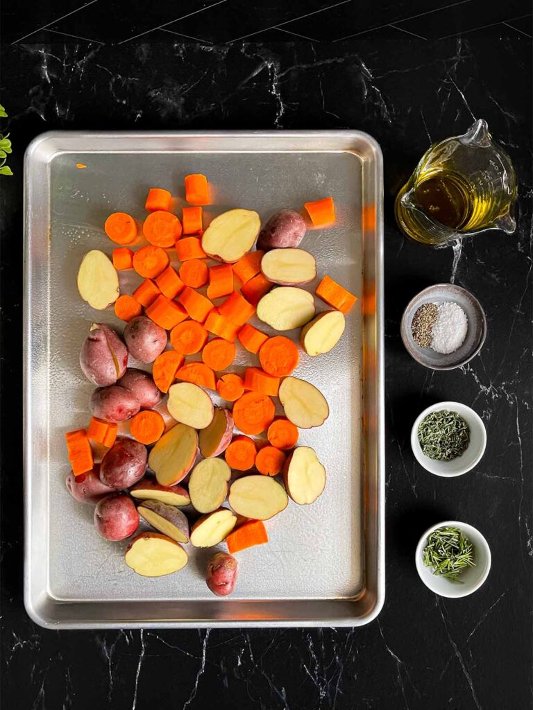 Potatoes and carrots on a baking sheet with the seasonings in ramekins next to the pan.
