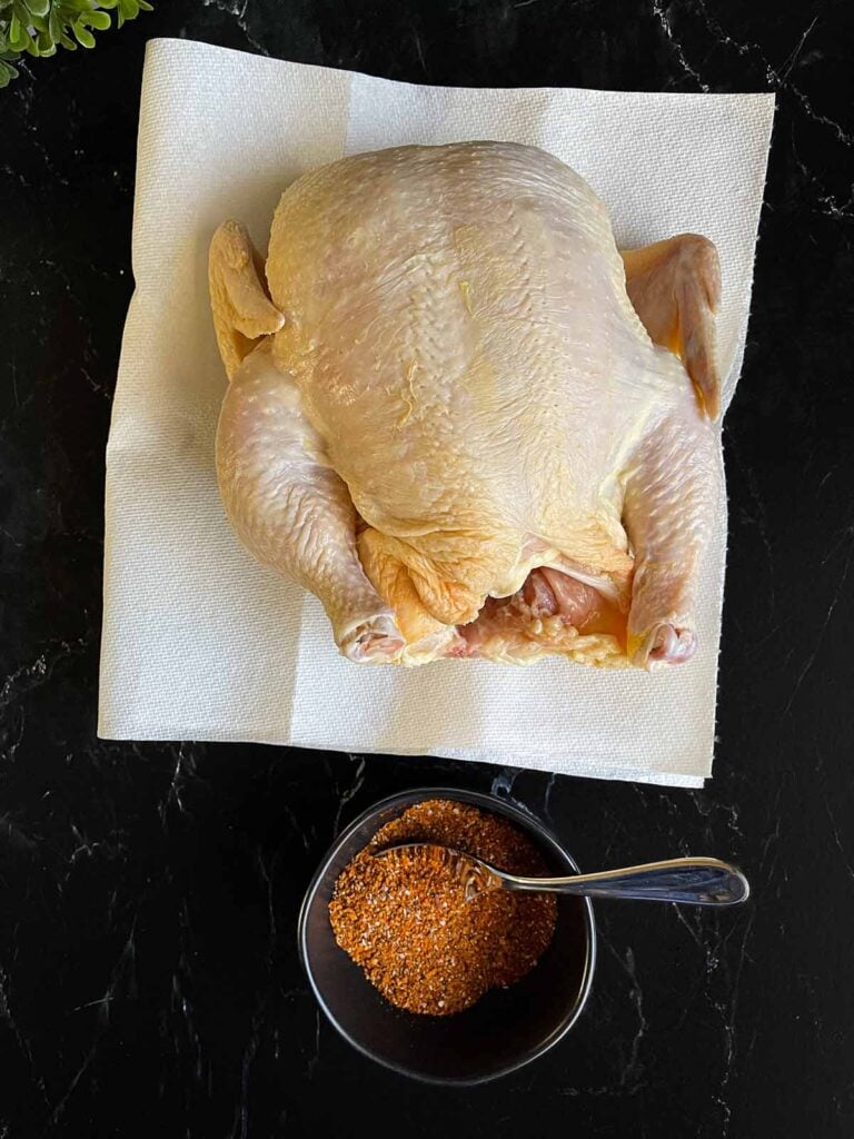 Raw whole chicken on paper towels and spice rub in a dark ramekin on a dark surface.