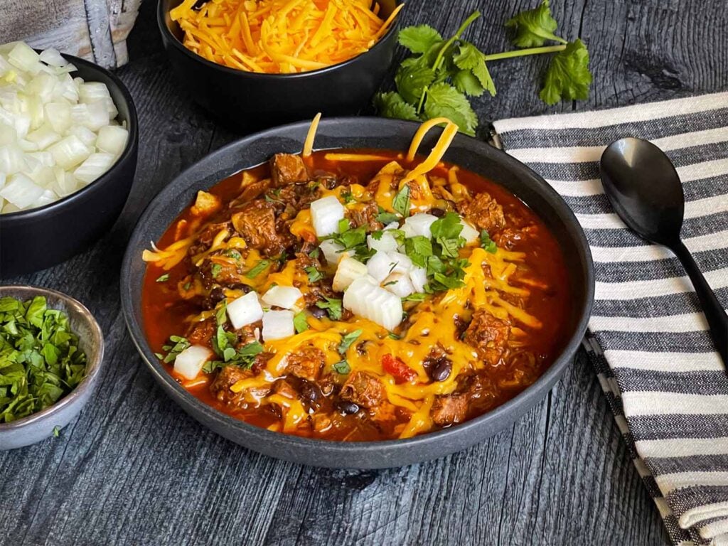 Brisket chili garnished with onions, cilantro, and cheddar cheese in a bowl sitting on a wooden background.