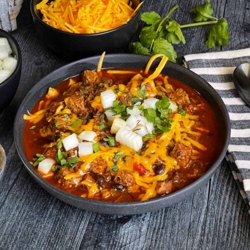 Brisket chili garnished with onions, cilantro, and cheddar cheese in a bowl sitting on a wooden background.