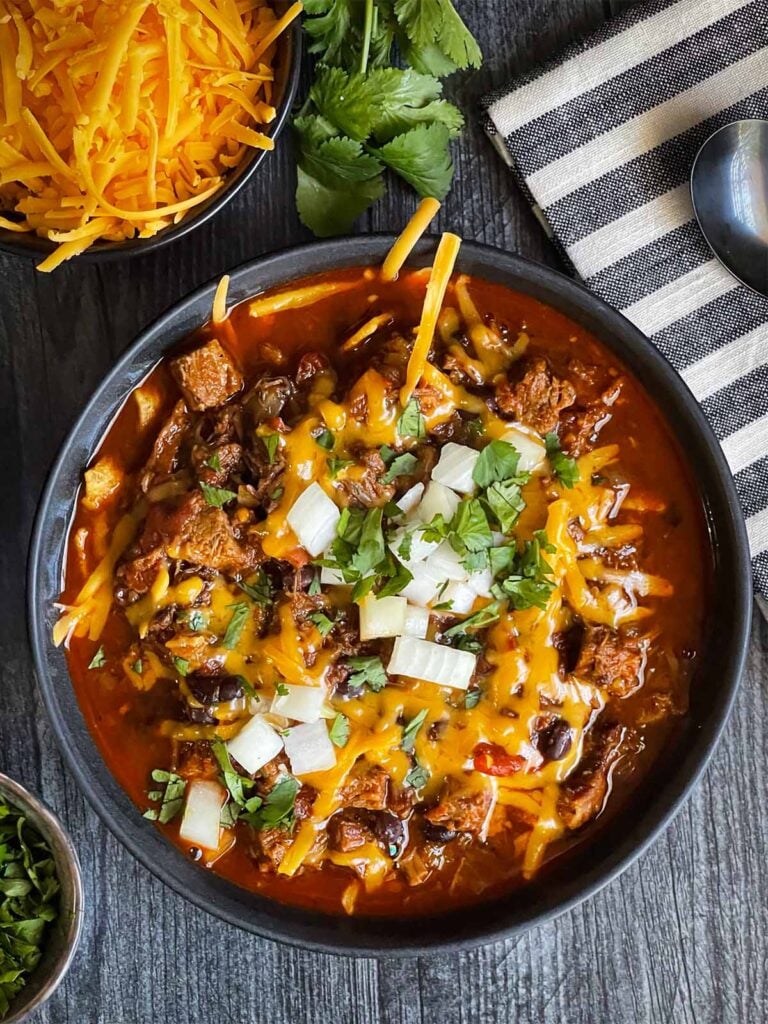 Brisket chili garnished with onions, cilantro, and cheddar cheese in a bowl sitting on a wooden background.