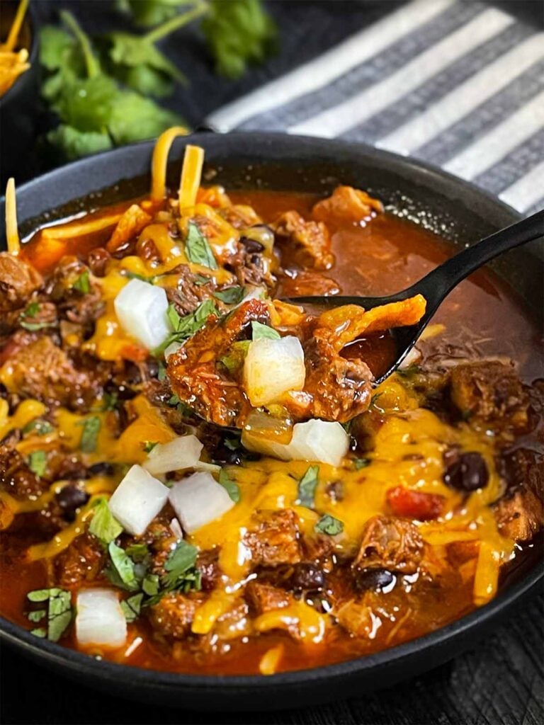 Brisket chili garnished with onions, cilantro, and cheddar cheese in a bowl sitting on a wooden background.