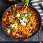 Brisket chili garnished with onions, cilantro, and cheddar cheese in a bowl sitting on a wooden background.