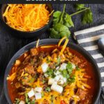 Brisket chili garnished with onions, cilantro, and cheddar cheese in a bowl sitting on a wooden background.