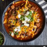 Brisket chili garnished with onions, cilantro, and cheddar cheese in a bowl sitting on a wooden background.