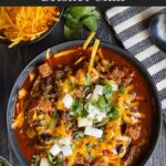 Brisket chili garnished with onions, cilantro, and cheddar cheese in a bowl sitting on a wooden background.