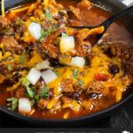 Brisket chili garnished with onions, cilantro, and cheddar cheese in a bowl sitting on a wooden background.