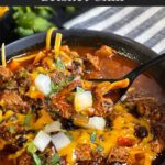 Brisket chili garnished with onions, cilantro, and cheddar cheese in a bowl sitting on a wooden background.