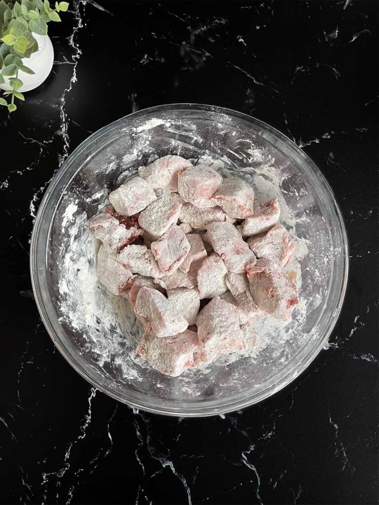 Seasoned beef tips dredged with flour in a glass mixing bowl on a dark surface.