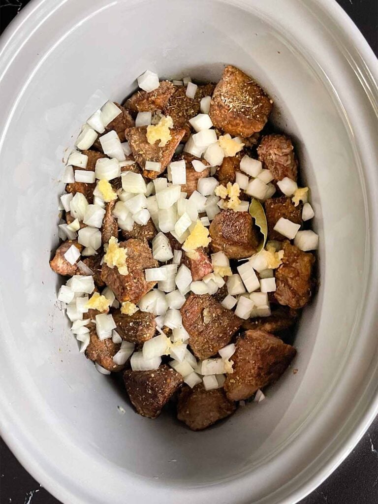Seasonings, onions, and garlic added to the beef tips in the crock of a slow cooker.