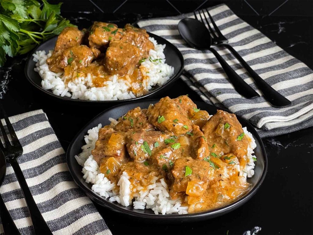 Slow cooker beef tips and gravy over cooked rice on a dark plate on a dark surface.