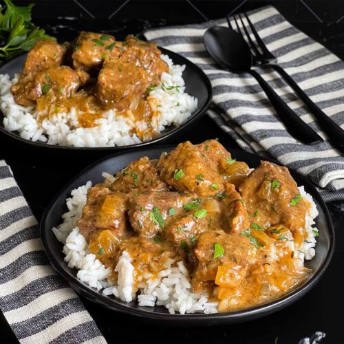 Slow cooker beef tips and gravy over cooked rice on a dark plate on a dark surface.