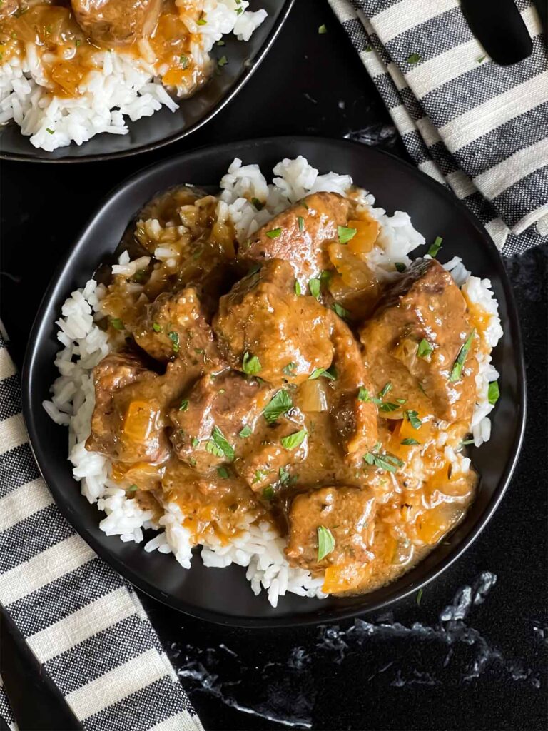 Slow cooker beef tips and gravy over cooked rice on a dark plate on a dark surface.